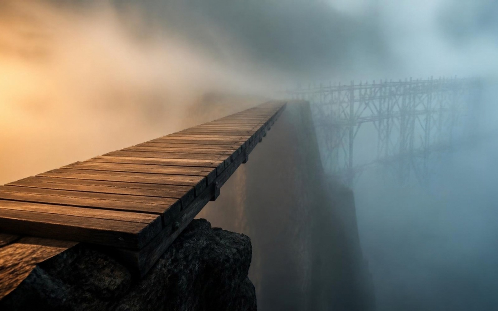 A wooden plank bridge extending from a rocky ledge into dense fog, with the silhouette of an unfinished scaffold barely visible on the far side