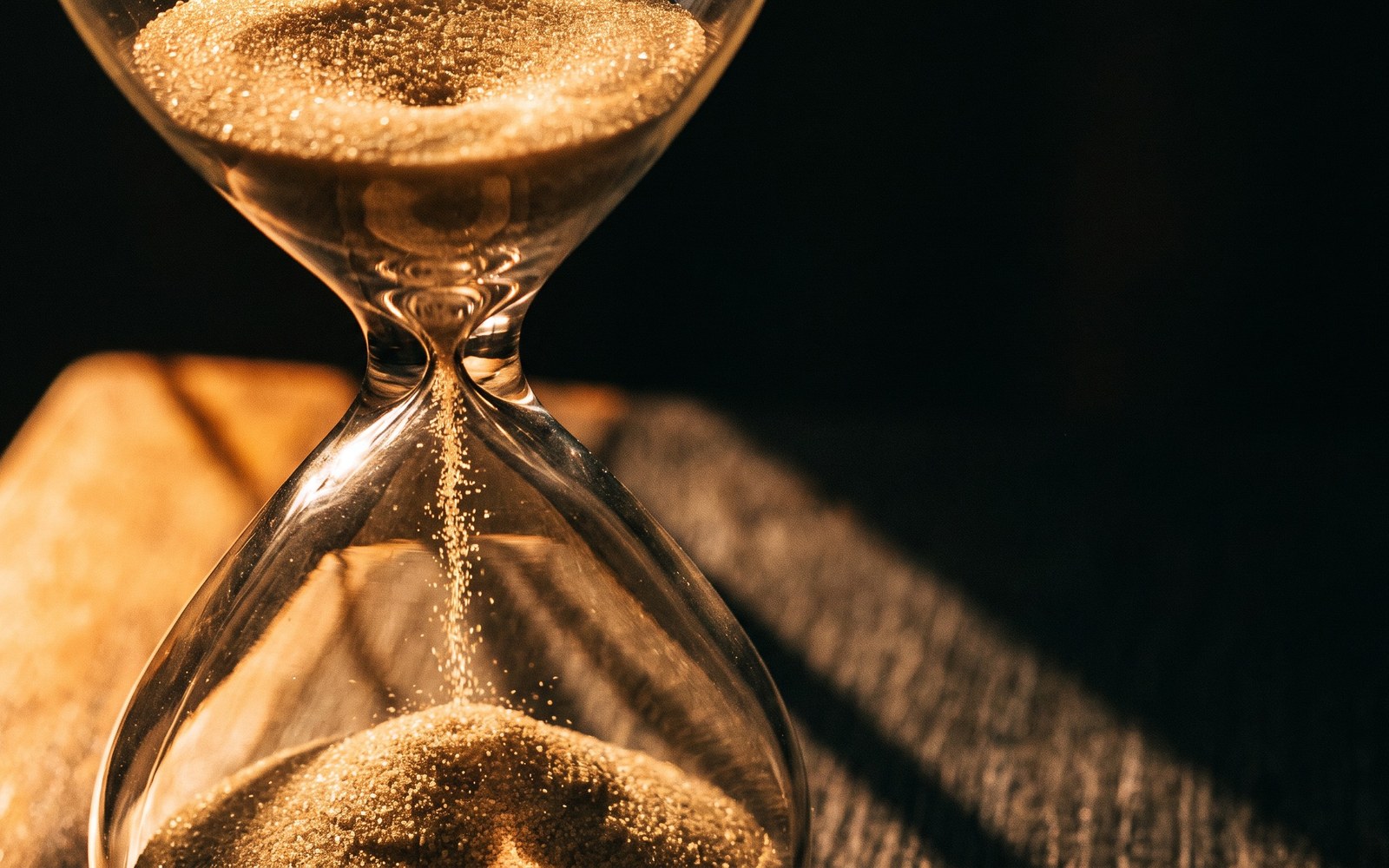 An hourglass mid-flow, sand falling in warm amber light against a dark background