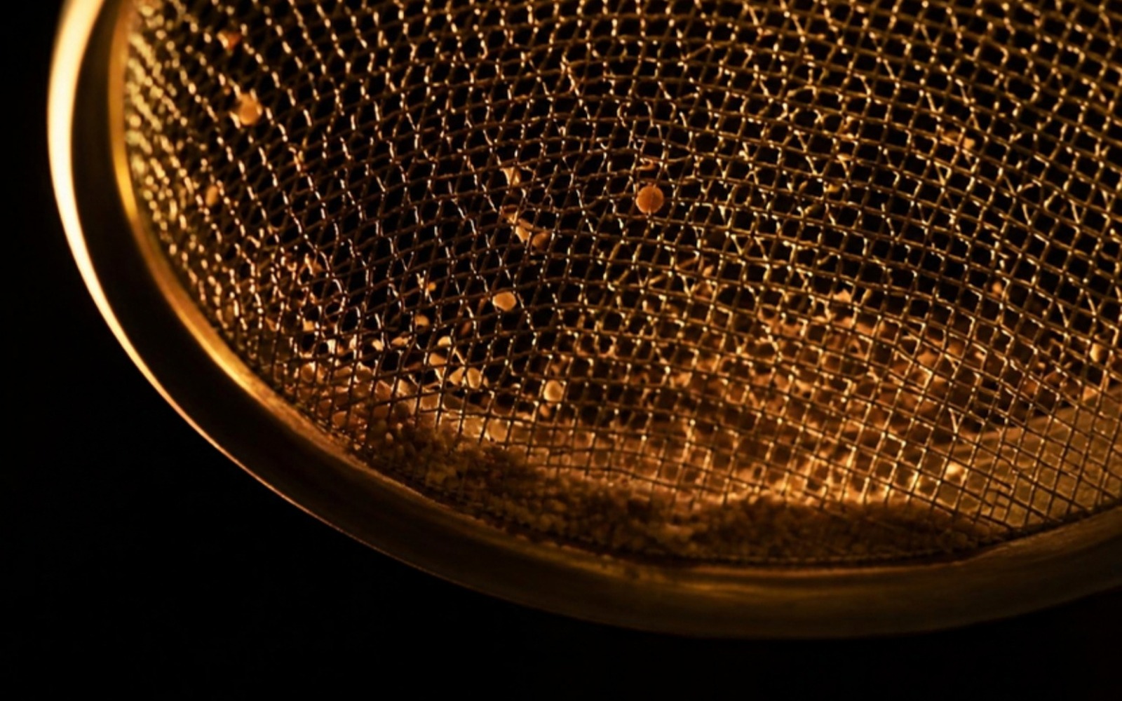 Macro photograph of a fine brass mesh sieve backlit with warm amber light, with a few grains caught in the mesh while most light passes through