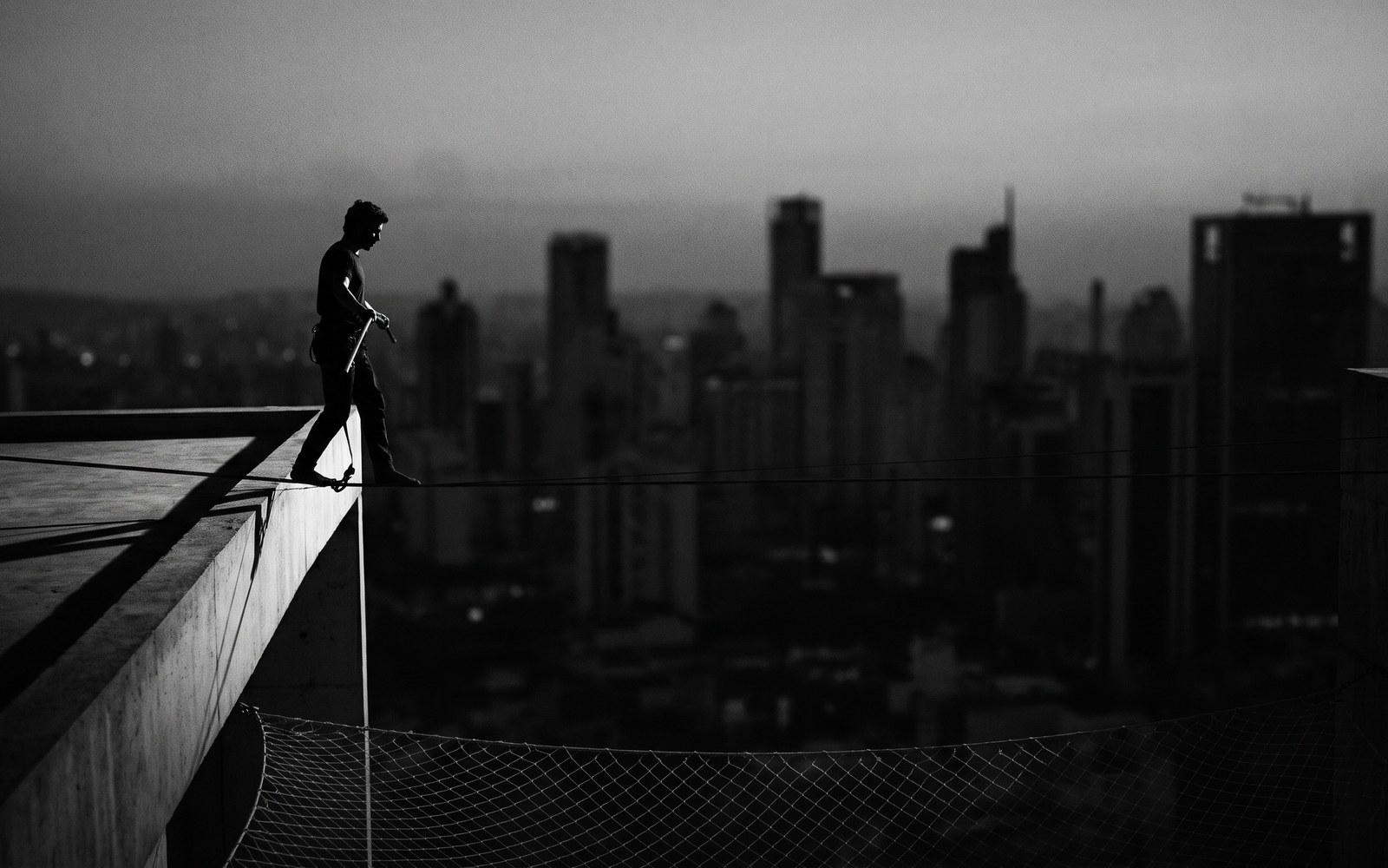 A lone tightrope walker crossing a wire high above a dim city at dusk, a faint safety net barely visible far below