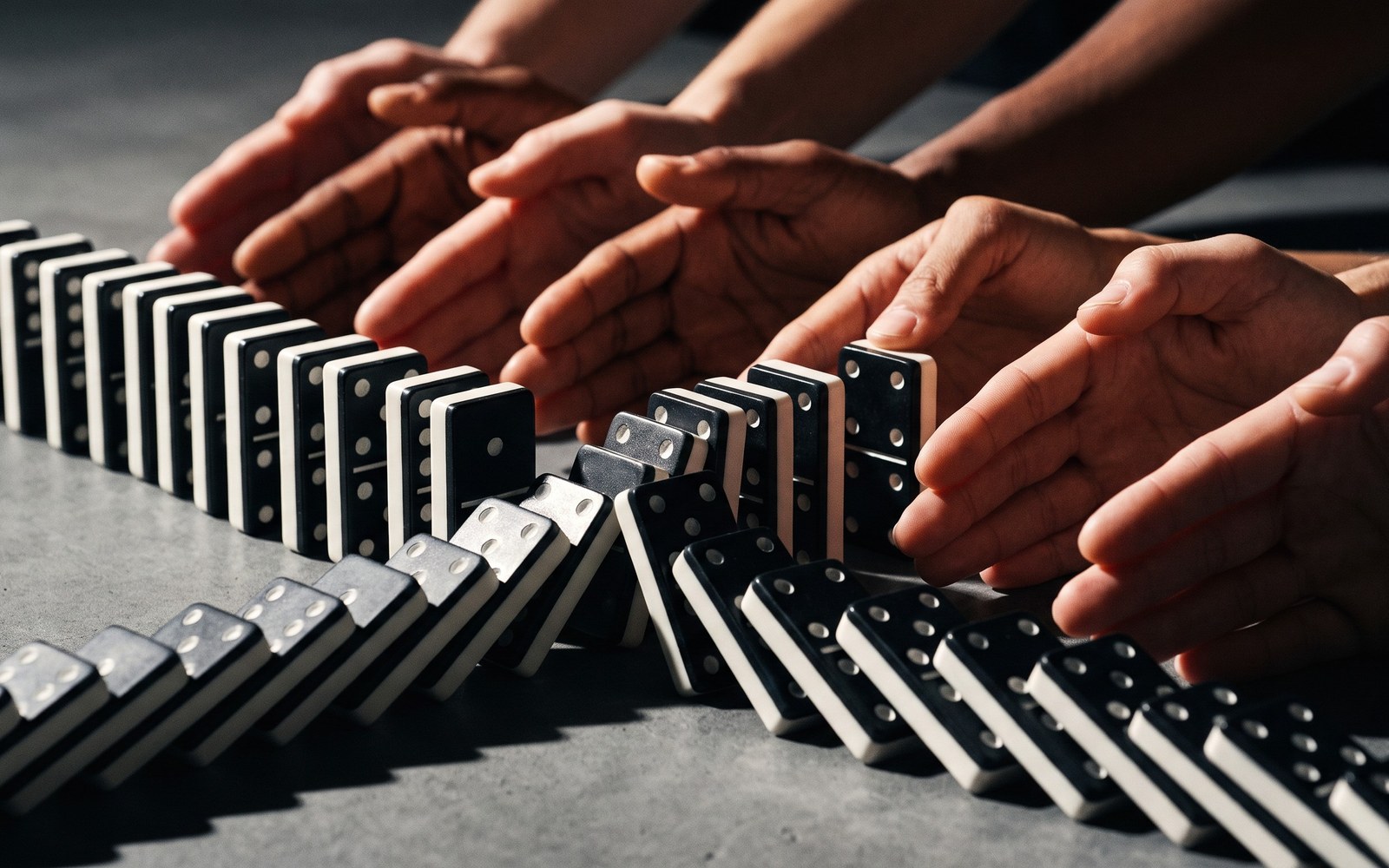 A chain of dominoes falling forward, with a second smaller chain of hands carefully standing each one back up in reverse order behind it