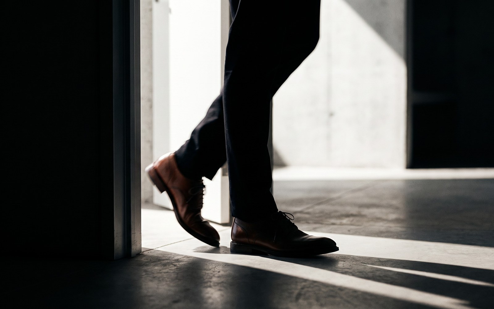 A person's silhouette paused mid-step in an open doorway between a bright sunlit room and a dim shadowed room