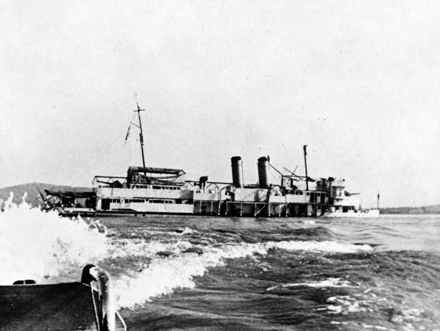 A black-and-white U.S. Navy photograph of USS Panay, a river gunboat, sinking in the Yangtze River after being attacked by Japanese aircraft, with a small motor sampan carrying survivors visible nearby.